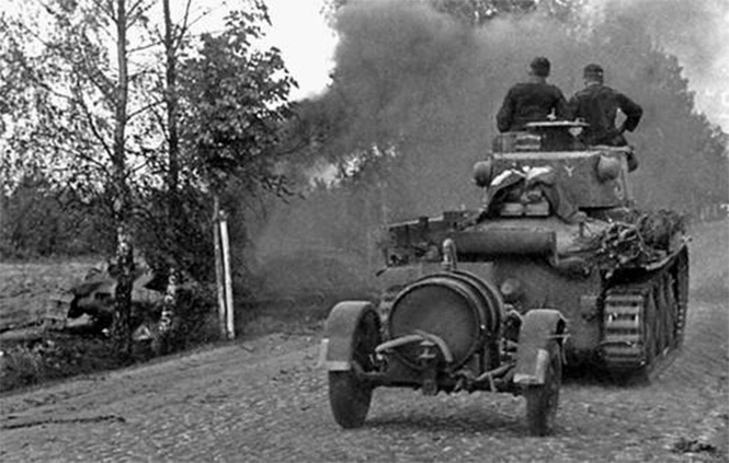 Немецкий танк PzKpfw38(t) едет мимо горящего советского танка. На буксире у «чеха» – бочка с горючим. Район Алитуса